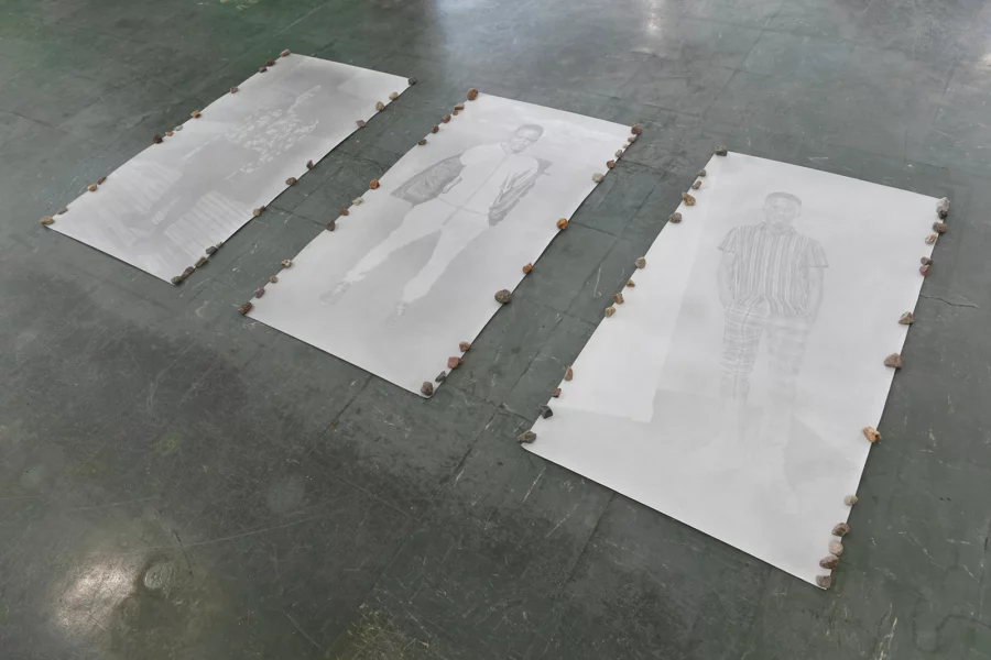 Aniruddha Biswas, En devenir, 2022–2023, installation view of large graphite portraits on paper placed on the floor and held down by small stones.