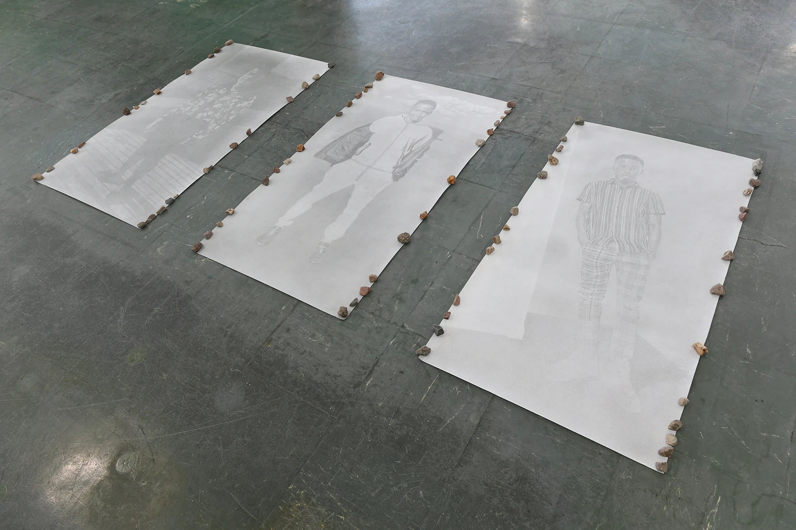 Aniruddha Biswas, En devenir, 2022–2023, installation view of large graphite portraits on paper placed on the floor and held down by small stones.